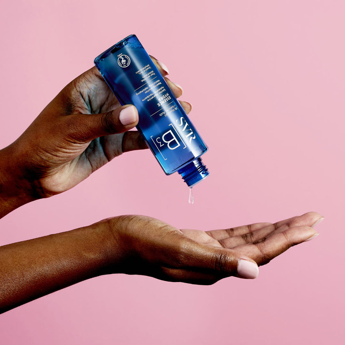 Flacon bleu SVR B3 Essence Hydra versant une goutte de soin liquide dans une main sur fond rose.