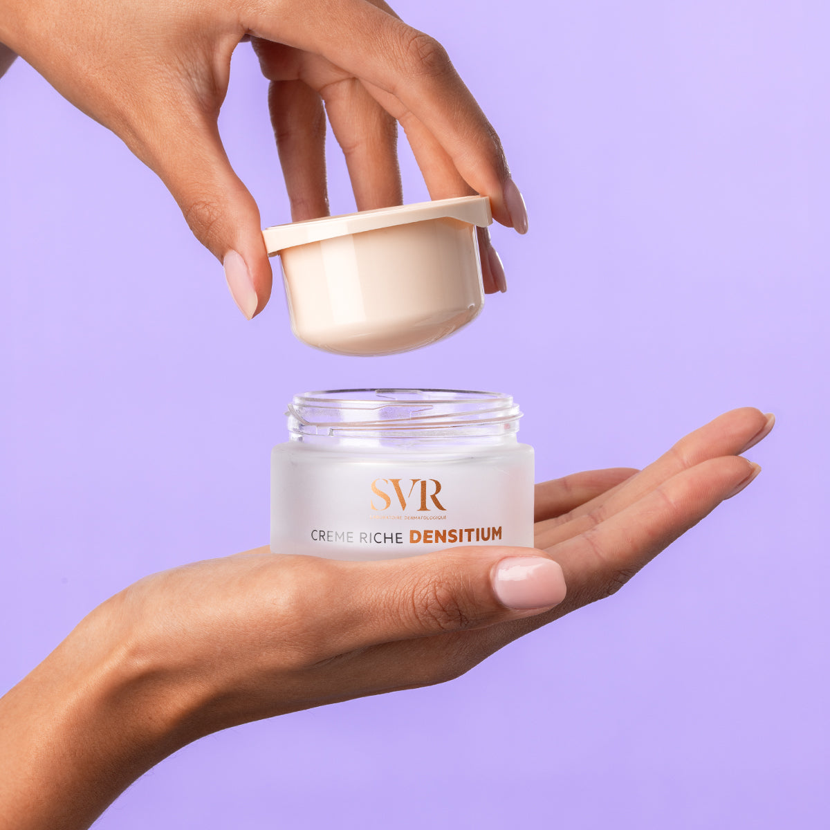 Hands holding an open SVR Densitium Crème Riche jar with a beige lid and frosted glass base against a purple background.