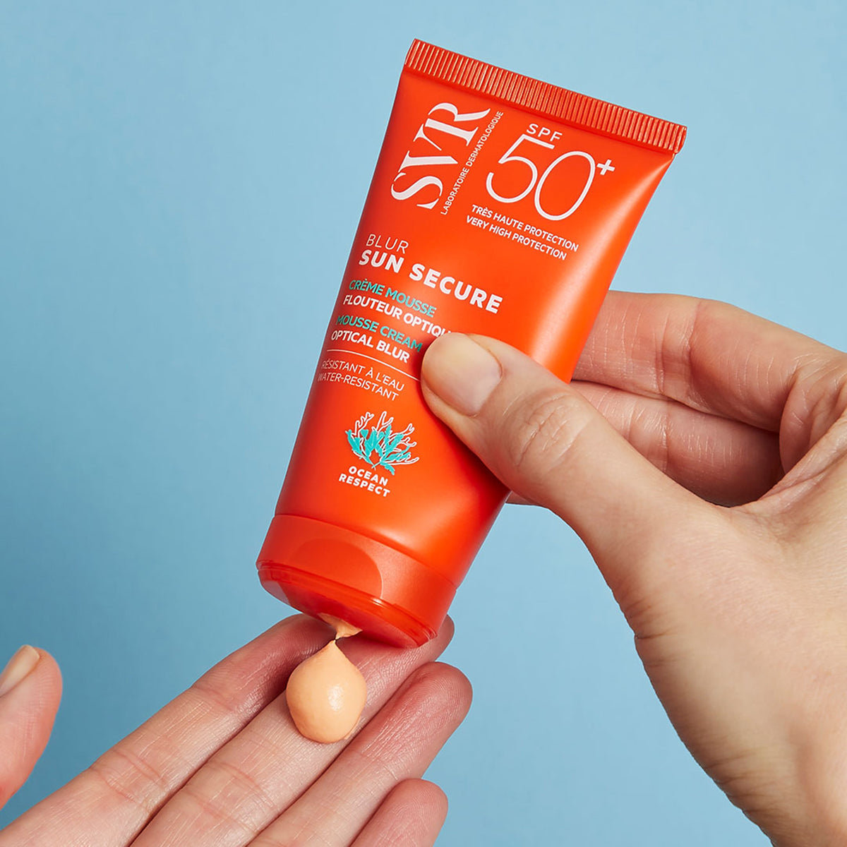 Tube orange SVR SUN SECURE Blur SPF50+ délivrant une crème mousse abricotée sur une main, sur fond bleu.