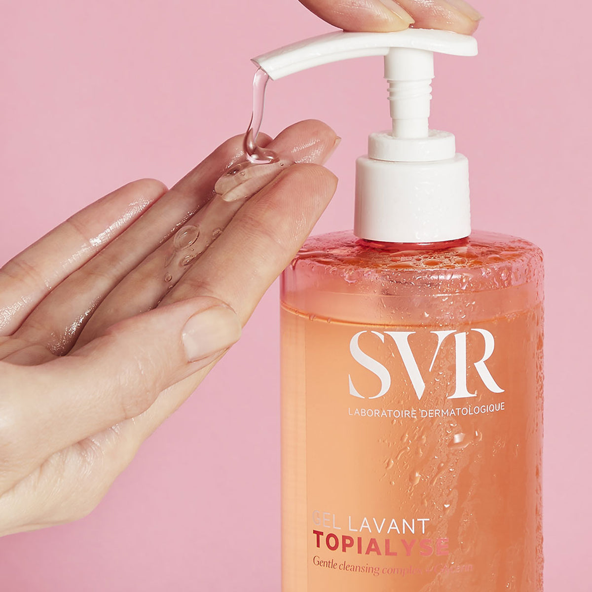Flacon pompe SVR Topialyse Gel Lavant transparent avec texture gel fluide versée dans une main sur fond rose.
