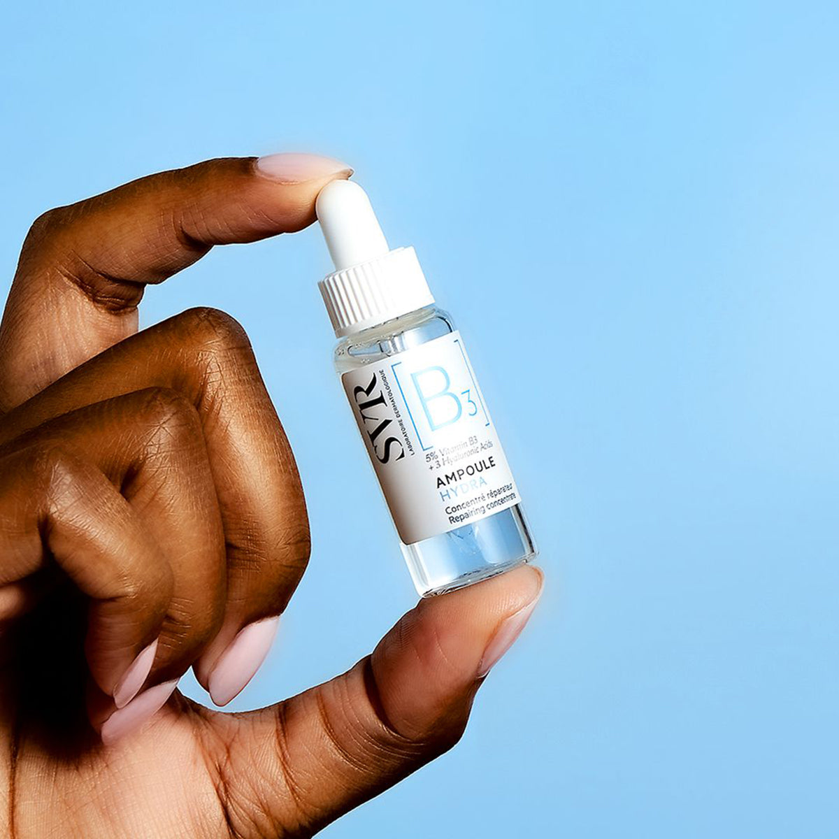 Main tenue par une main, flacon compte-gouttes transparent SVR Ampoule Hydra avec étiquette blanche sur fond bleu.