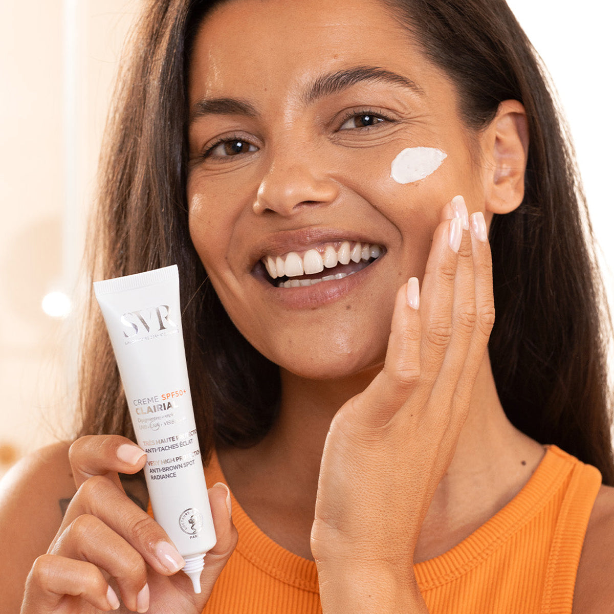 Femme souriante appliquant la crème solaire dépigmentante SVR Clairial SPF50+ sur son visage.
