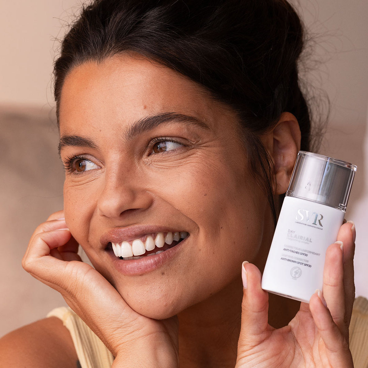 Femme souriante tenant un flacon blanc SVR Clairial Day anti-taches avec bouchon transparent.