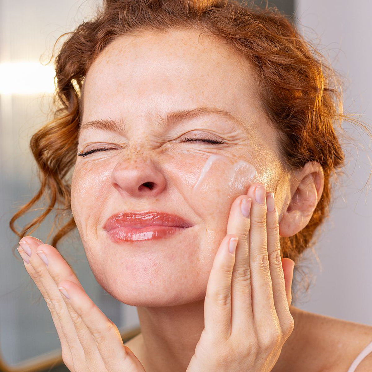Femme rousse aux taches de rousseur appliquant le baume démaquillant SVR Sensifine sur son visage avec ses mains.