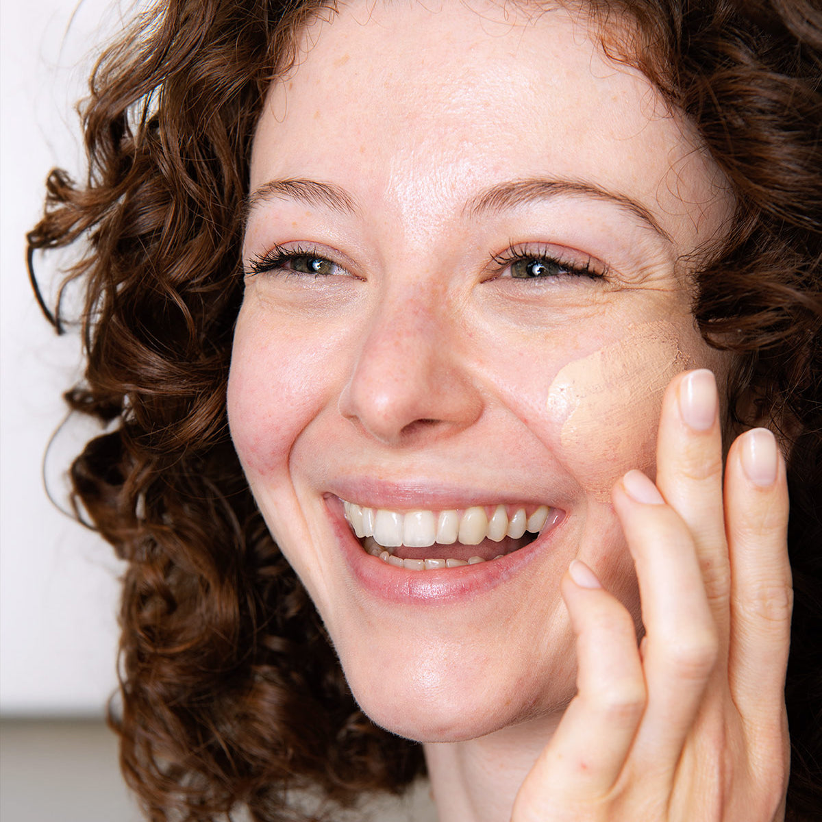 Femme souriante aux cheveux bouclés appliquant la crème teintée SVR Sensifine AR sur sa joue pour camoufler les rougeurs.