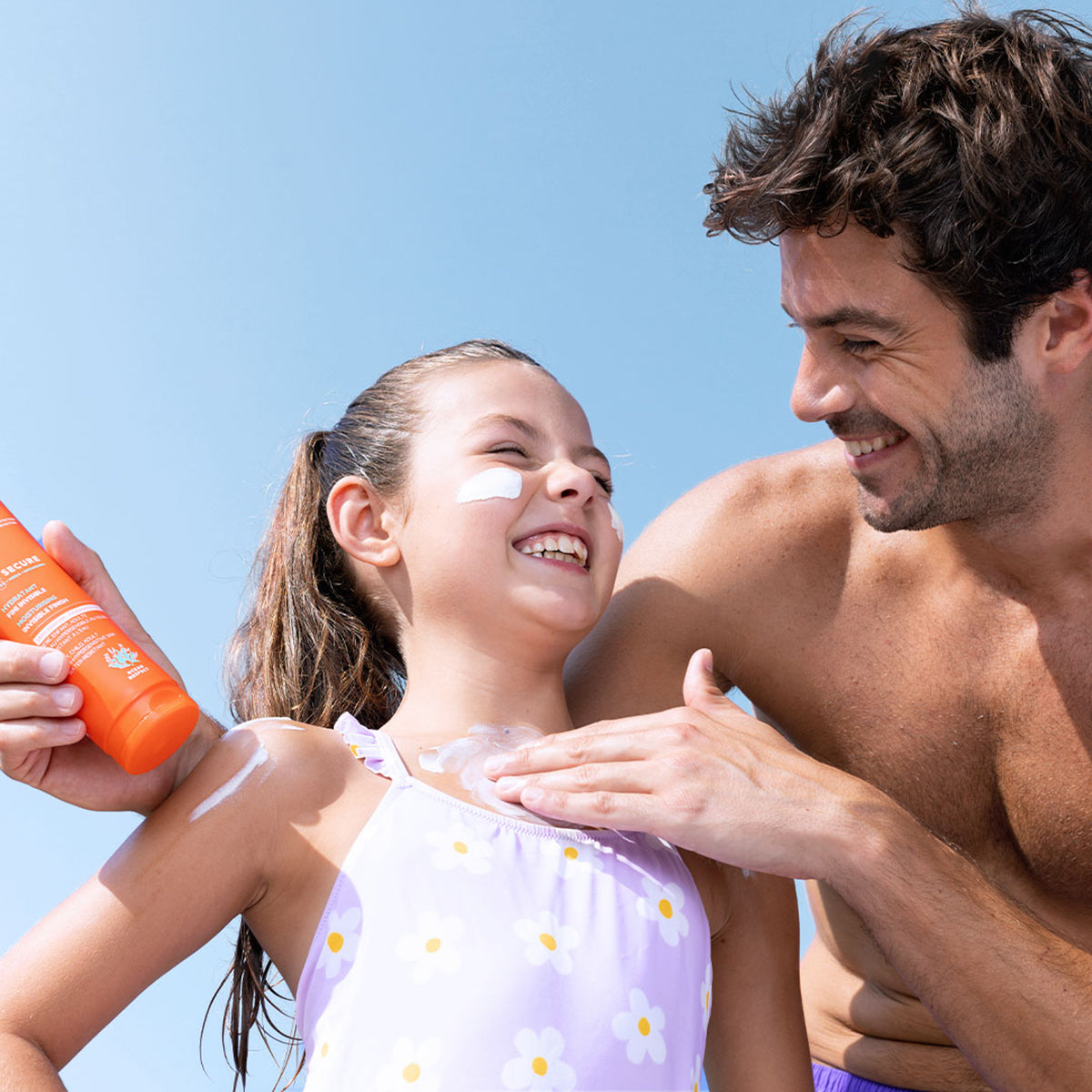 Père appliquant le lait solaire orange SUN SECURE SPF50+ sur les épaules de sa fille souriante sous un ciel bleu.