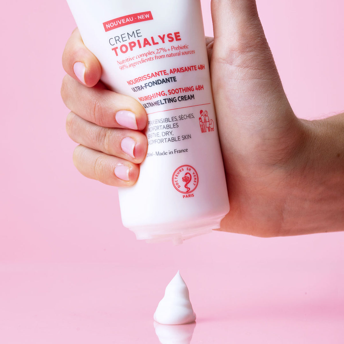 Main tenue pressant un tube blanc de crème hydratante TOPIALYSE sur fond rose, avec une noisette de produit onctueux.