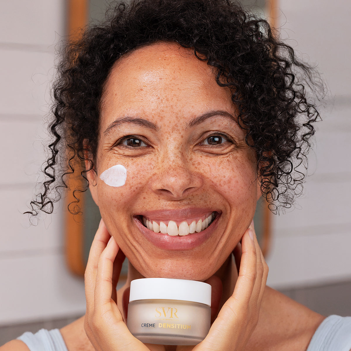 Smiling woman with curly hair holding a jar of SVR Densitium Creme with a dab of white face cream on her cheek.