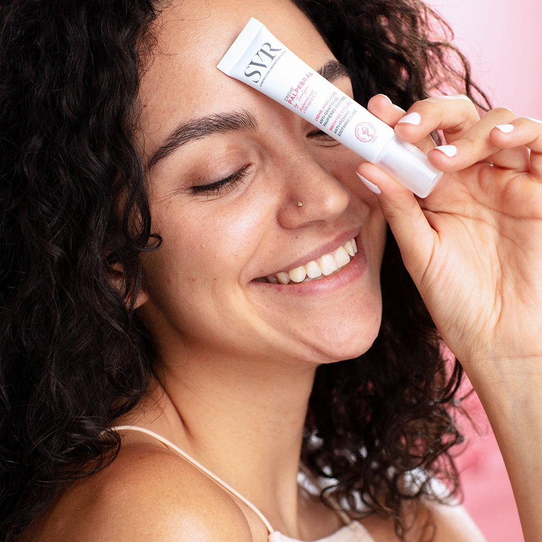 Femme souriante tenant un tube blanc de crème apaisante SVR Palpebral contre son front sur fond rose.