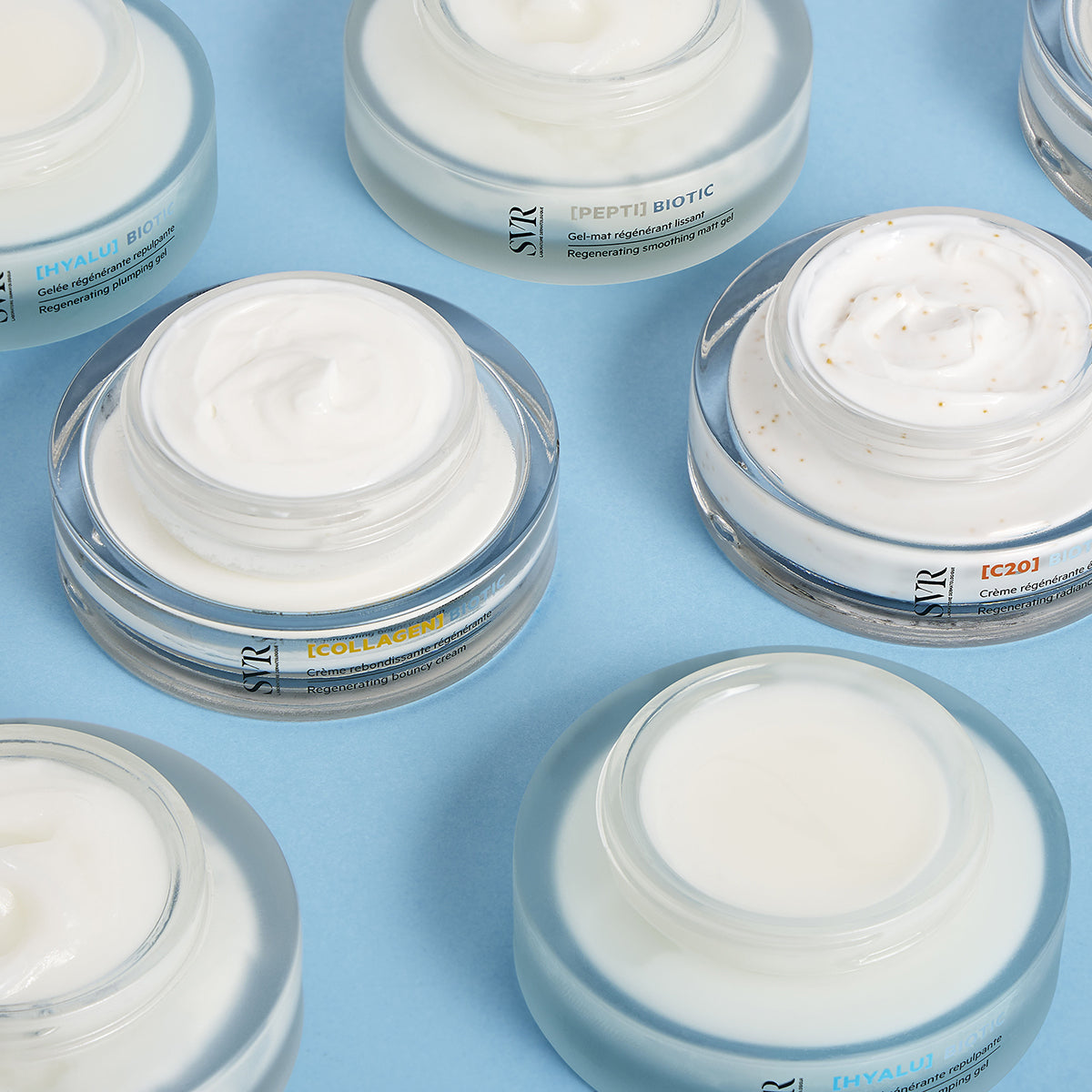 Pots de crème SVR Biotic ouverts avec textures onctueuses sur fond bleu, présentant les gammes Hyalu, Pepti, Collagen et C20.