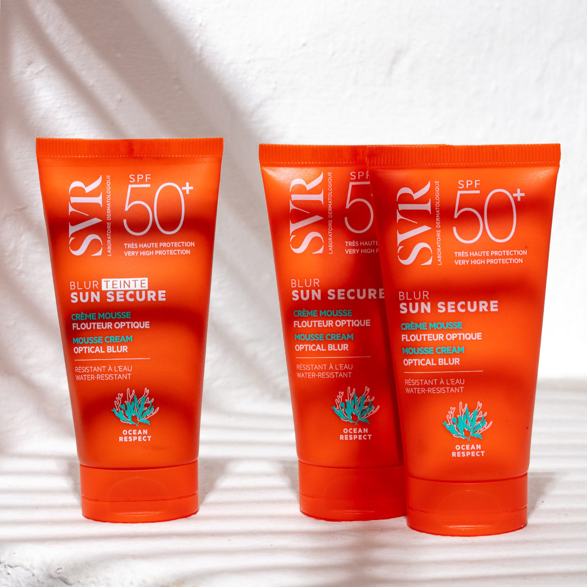 Trois tubes orange de crème solaire SVR Sun Secure Blur teinté SPF50+ sur fond blanc avec des ombres de feuilles.