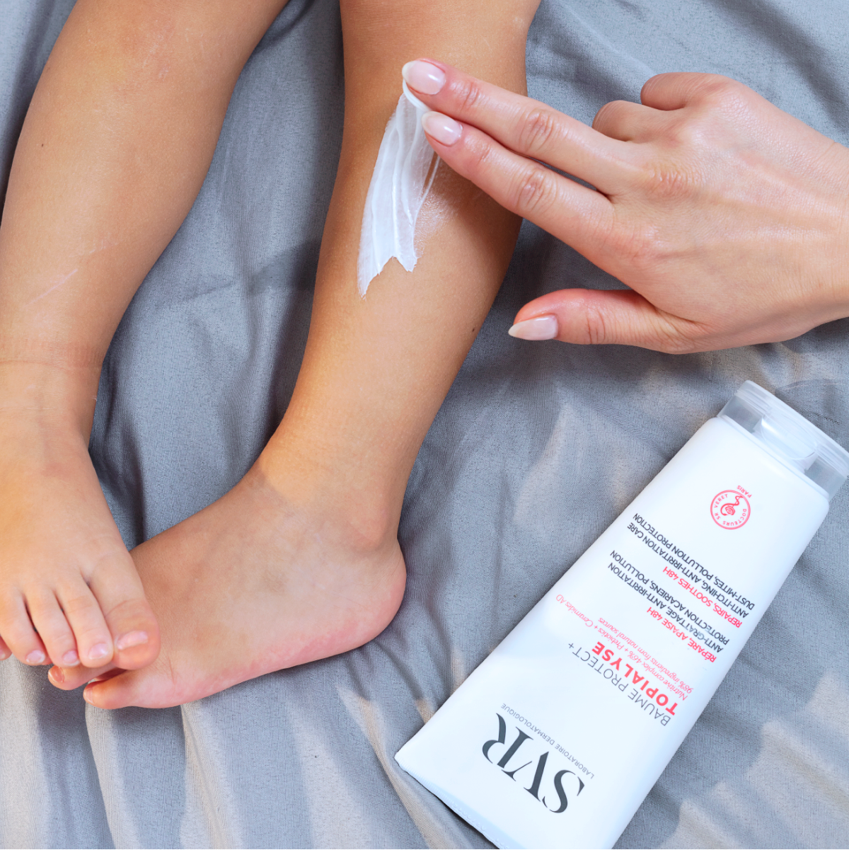 Main appliquée sur la jambe d'un enfant pour appliquer la crème apaisante SVR Topialyse sur un drap gris.