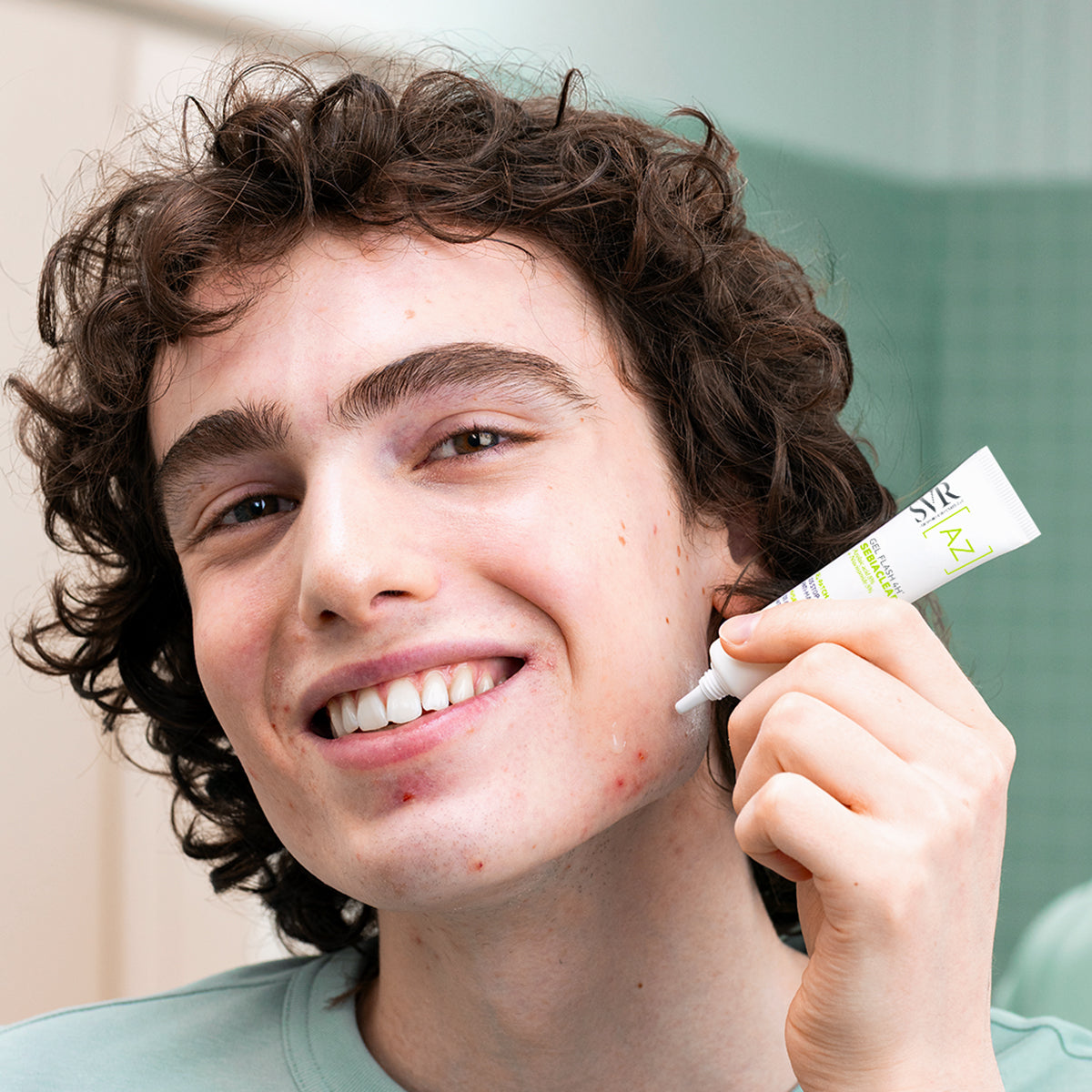 Young man applying SVR Sebiaclear Gel Flash 4H from a white squeeze tube onto a blemish on his cheek.