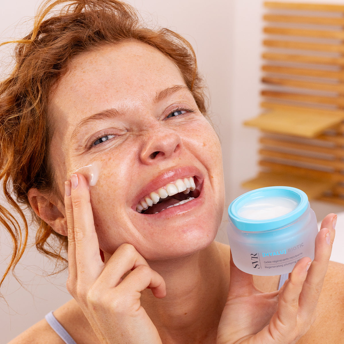 Smiling woman applying SVR HYALU Biotic regenerating plumping gel from a frosted glass jar with a blue lid.