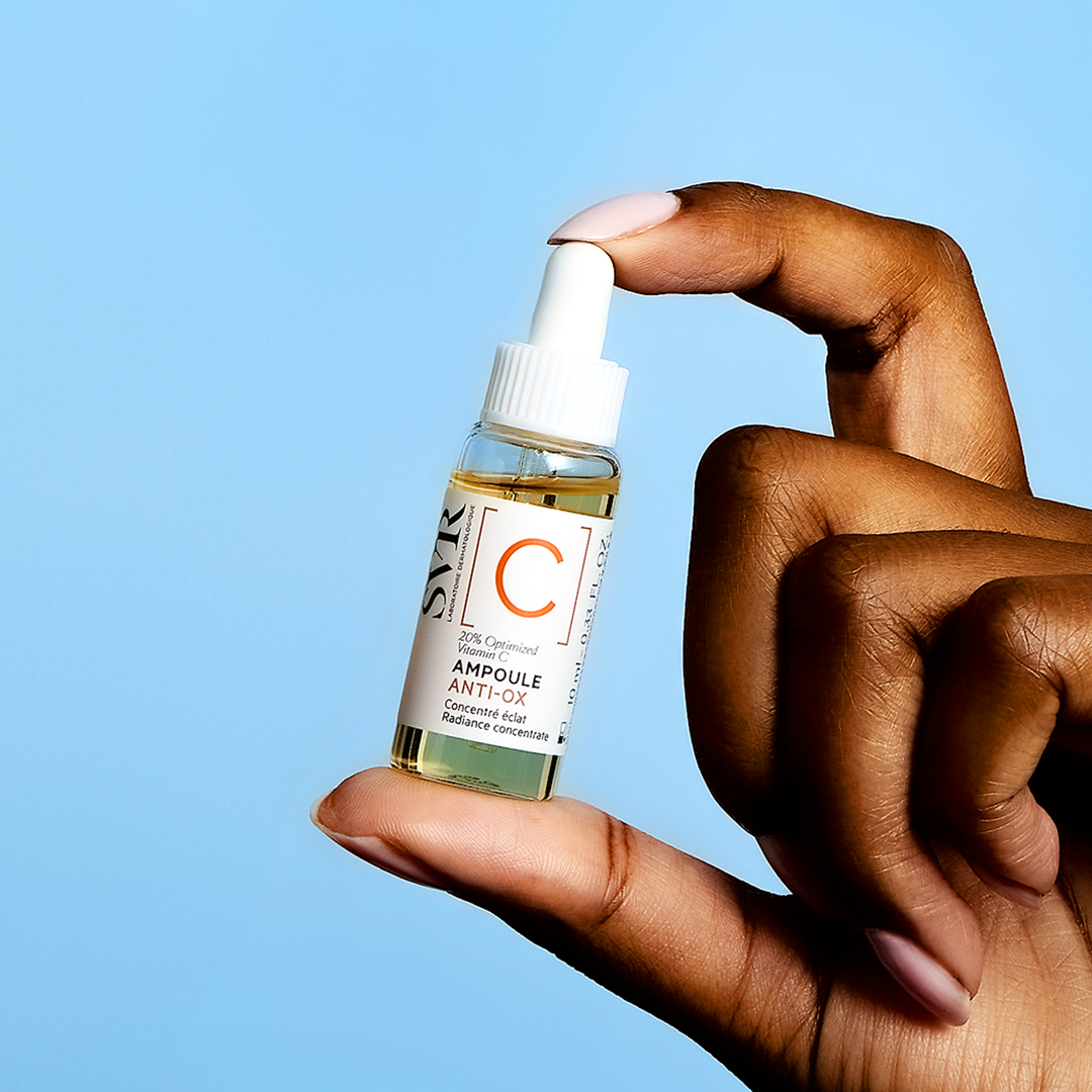 Main tenue entre les doigts, flacon compte-gouttes SVR Ampoule Anti-Ox de 10 ml avec sérum à la vitamine C sur fond bleu.