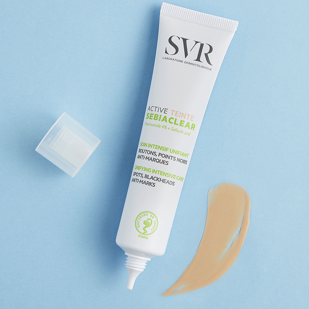 Tube de soin teinté SVR Sebiaclear Active avec bouchon ouvert et échantillon de crème beige sur fond bleu clair.