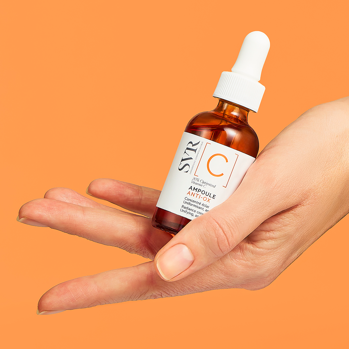 Main tenue dans la main, flacon compte-gouttes en verre ambré SVR Ampoule Anti-Ox sur fond orange vif.