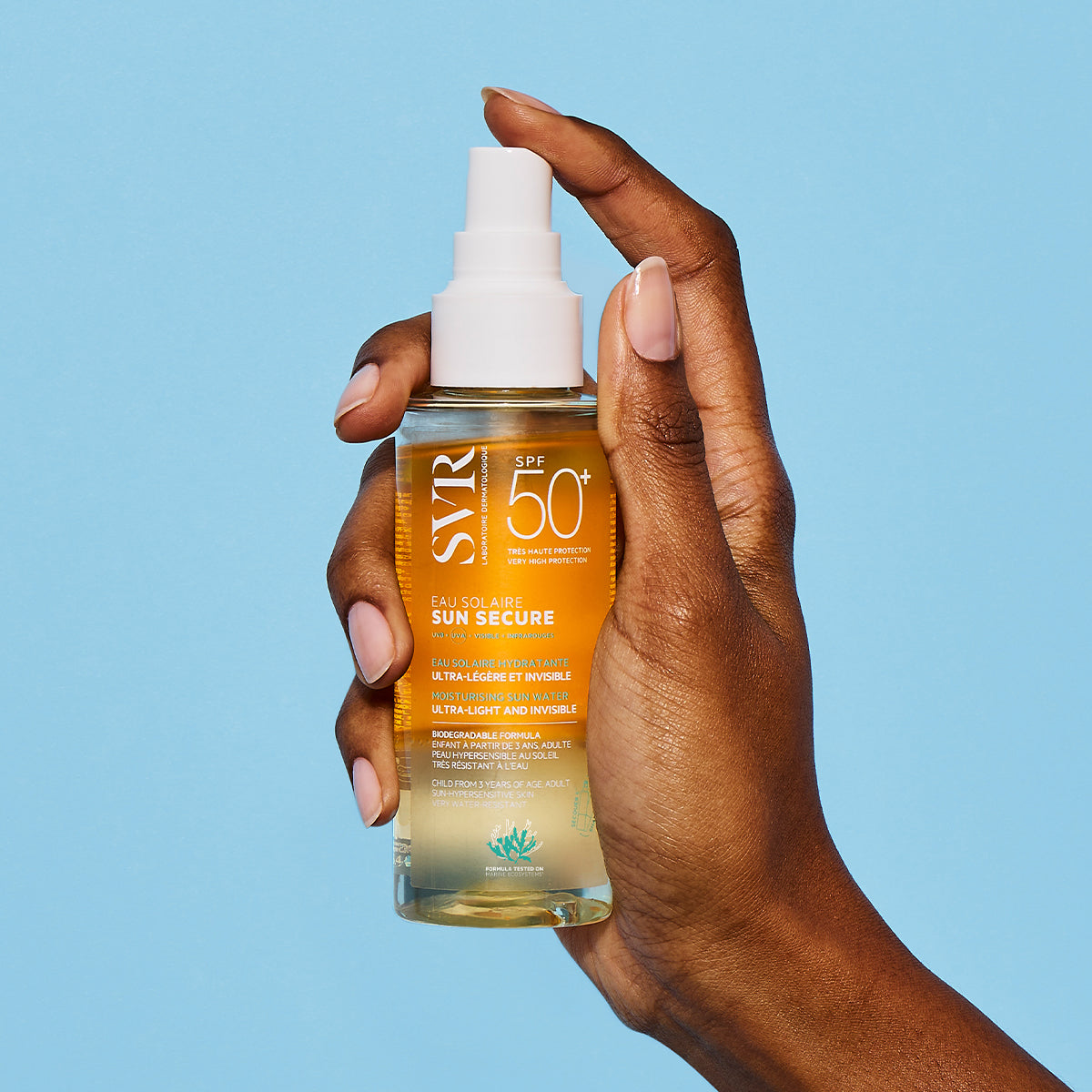 Main tenue par une main, flacon spray SVR Sun Secure Eau Solaire SPF50+ à la texture bi-phasée dorée sur fond bleu.