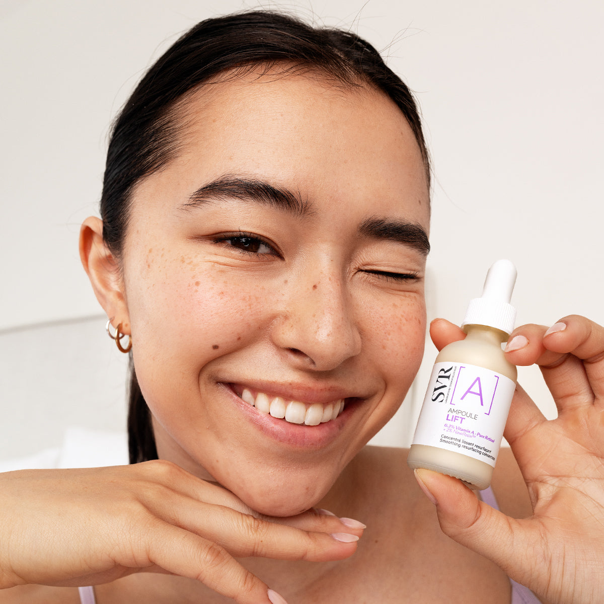 Smiling woman holding a small glass bottle of SVR Ampoule Lift serum with a white dropper cap and purple label.
