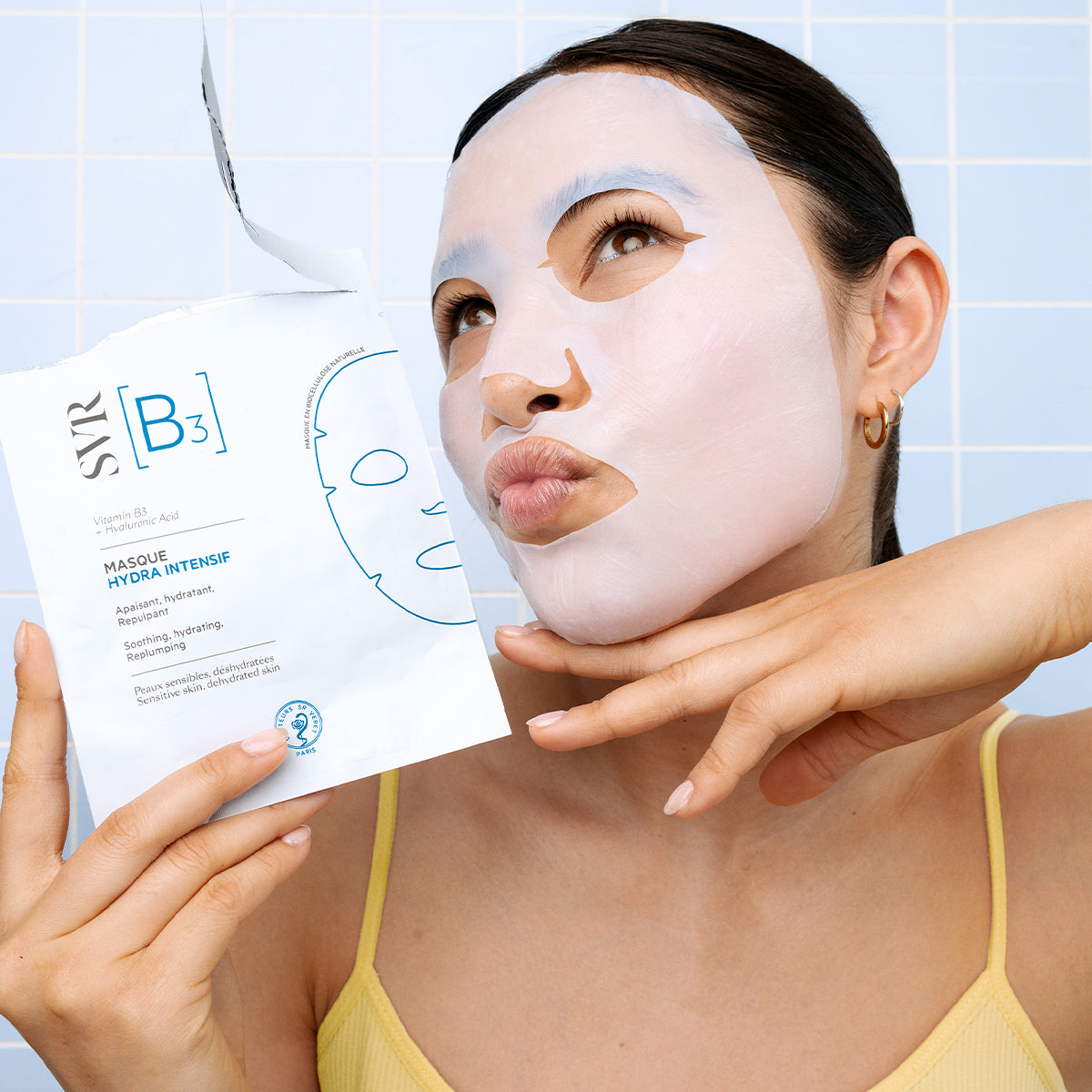 Femme portant un masque en biocellulose SVR B3 Hydra Intensif blanc devant un carrelage bleu clair.