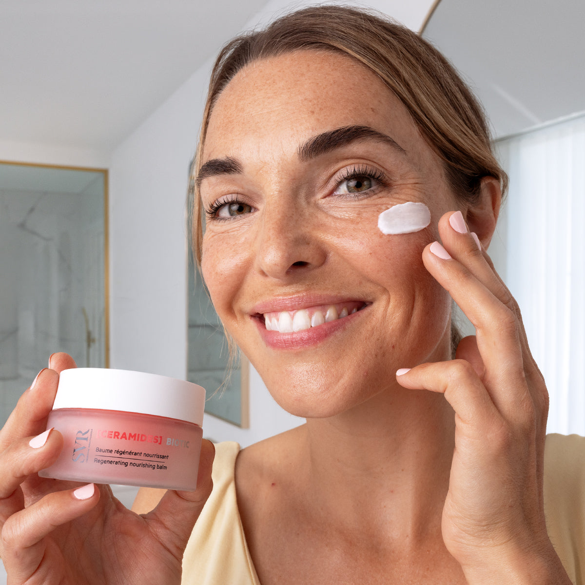 Woman smiling while applying SVR Ceramides Biotic regenerating nourishing balm from a pink glass jar to her cheek.