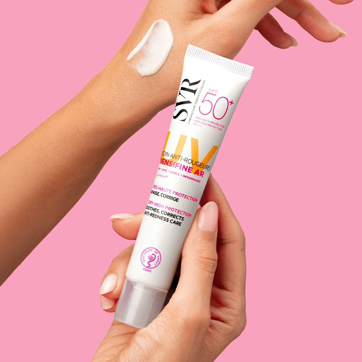 Tube de crème SVR Sensifine AR SPF50+ tenu en main avec une noisette de texture blanche sur le bras, sur fond rose.