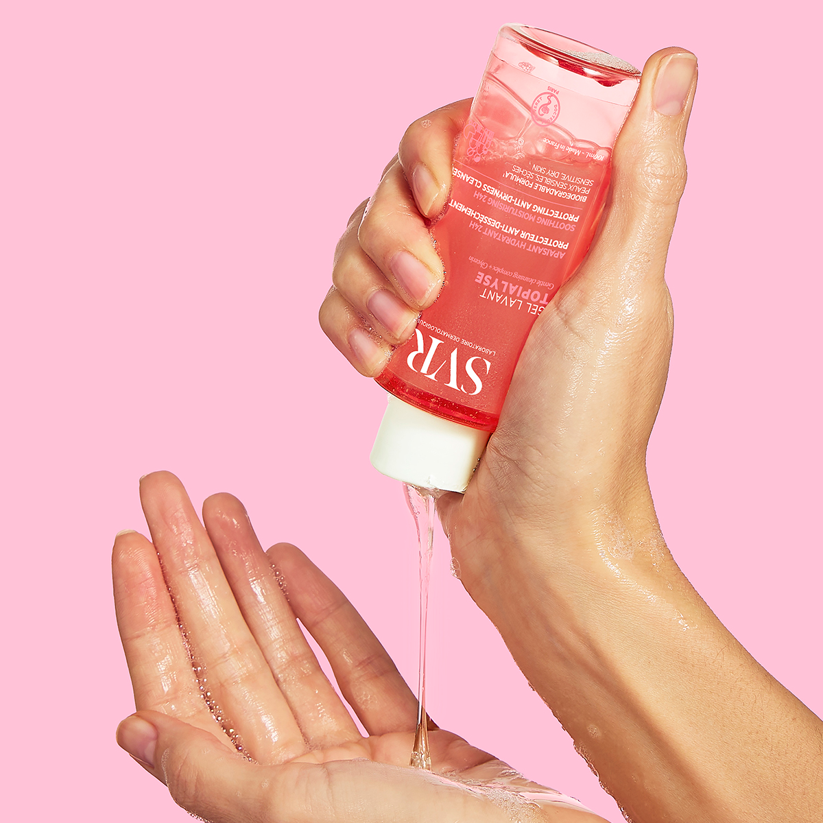 Main versant le gel lavant transparent SVR Topialyse depuis un flacon rose sur un fond rose pastel.