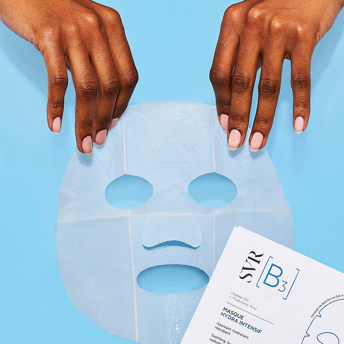Mains tenant un masque en biocellulose hydratant SVR B3 sur fond bleu, avec l'emballage du produit visible en bas.