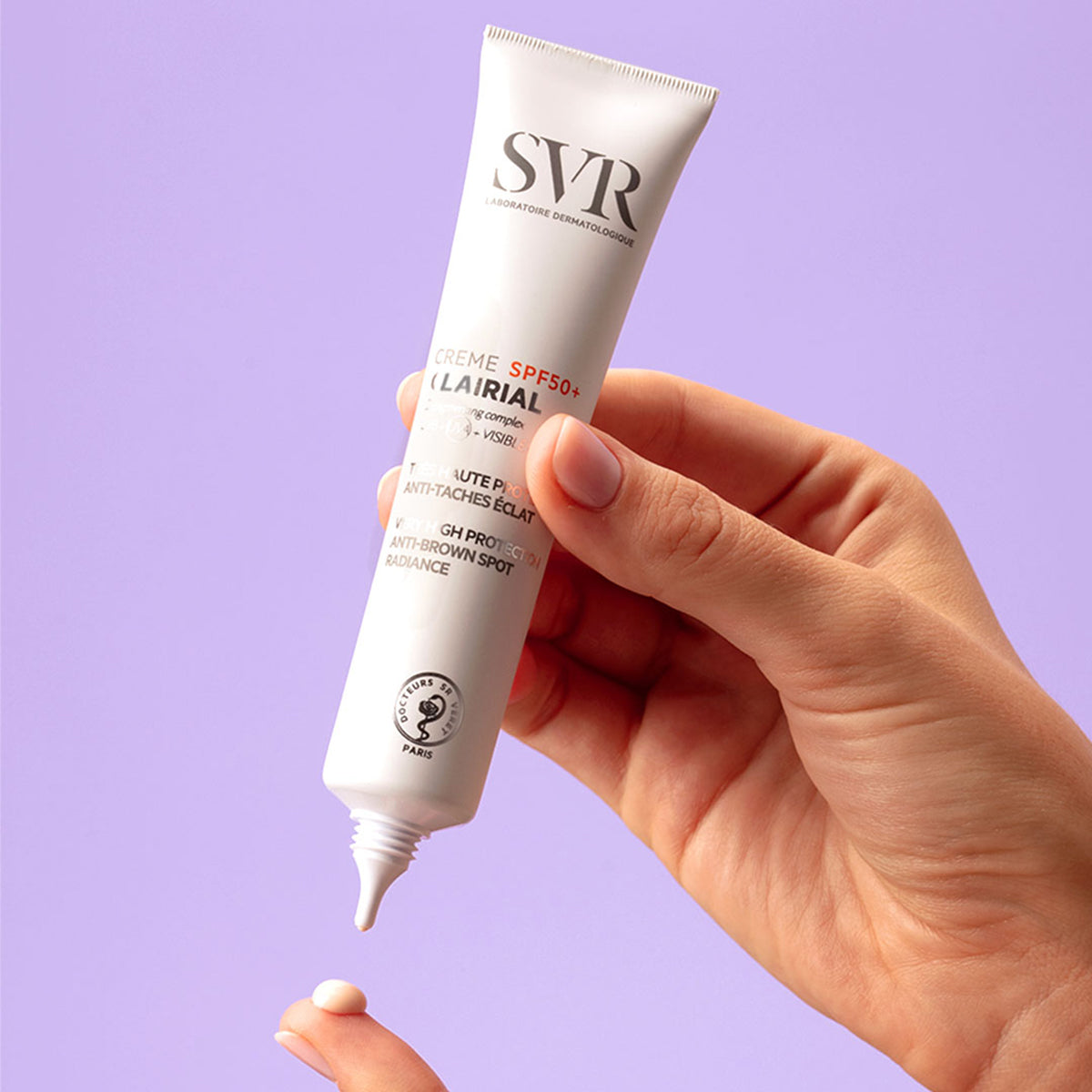 Main tenue en main pressant un tube blanc SVR Clairial Crème SPF50+ sur un fond violet pastel.
