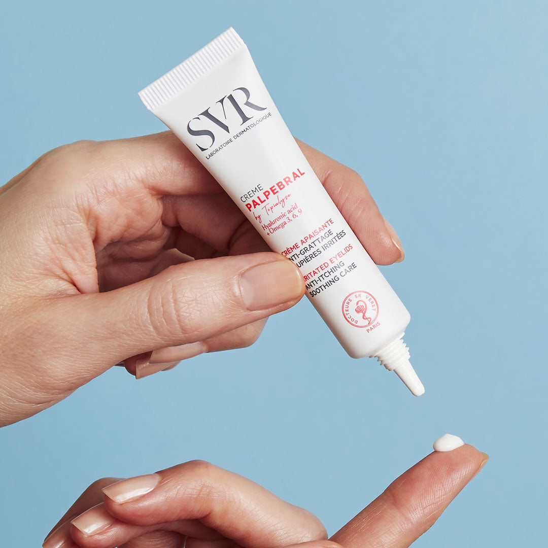 Tube blanc de crème SVR Palpebral pressé au-dessus d'un doigt, texture onctueuse pour paupières sensibles sur fond bleu.