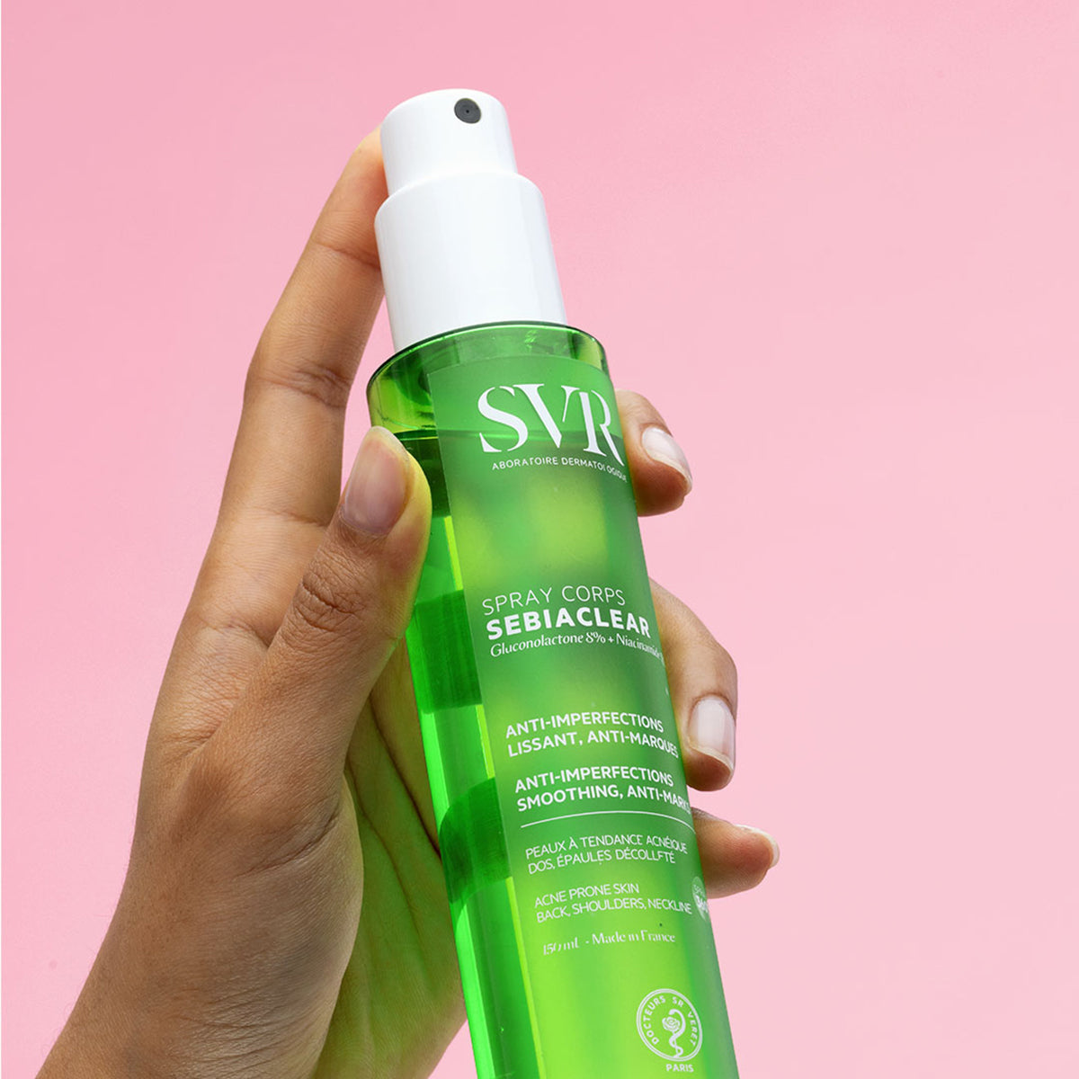 Main tenue par une main, flacon spray vert SVR Sebiaclear pour le corps sur fond rose.