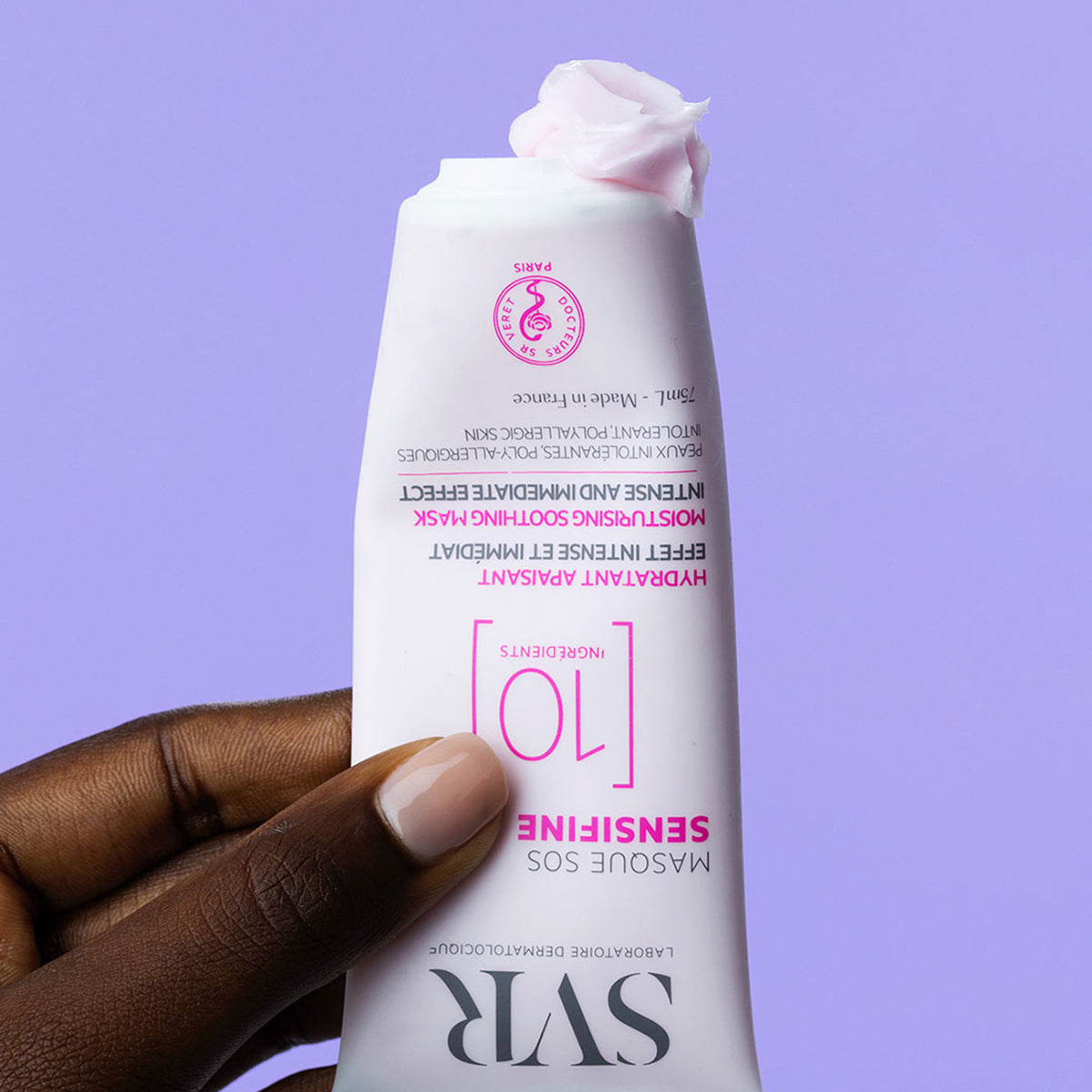 Tube de masque SVR Sensifine SOS rose avec texture crémeuse sortant de l'embout, tenu par une main sur fond violet.