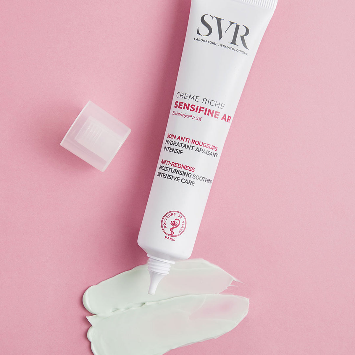 Tube blanc de crème SVR Sensifine AR avec bouchon ouvert et texture crème verte sur fond rose.