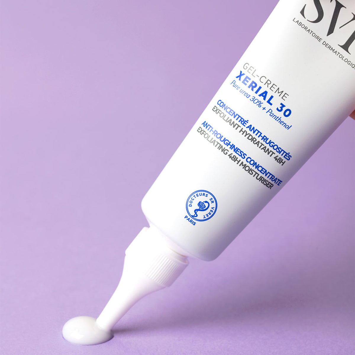 Tube blanc de SVR XERIAL 30 Gel-Crème déposant une goutte de crème exfoliante sur un fond violet pastel.