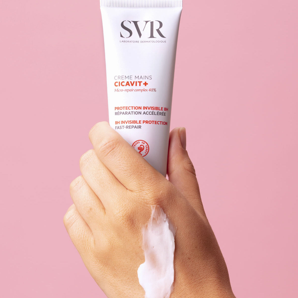 Main tenant un tube blanc de crème réparatrice SVR Cicavit+ sur fond rose, avec une texture crémeuse appliquée sur la peau.