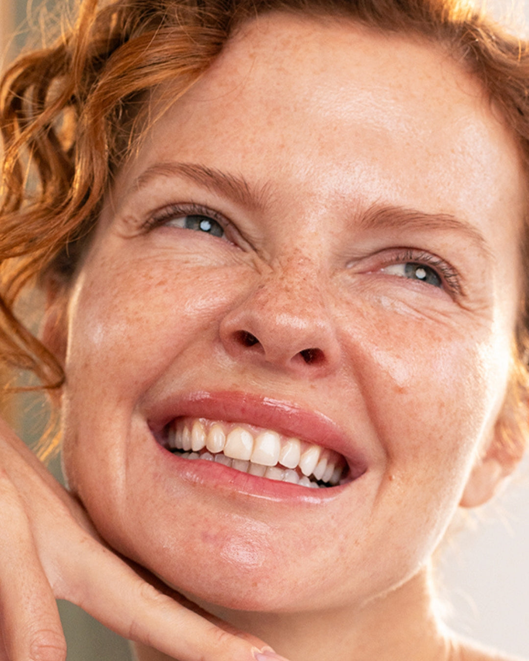 Close-up of a smiling woman with red curly hair, freckled skin, and a glowing, radiant complexion.