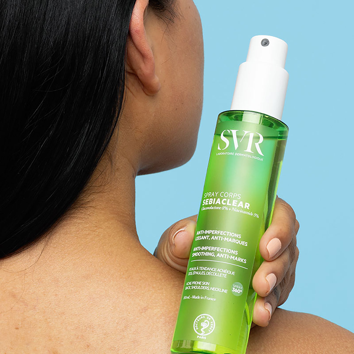 Flacon spray vert SVR Sebiaclear Corps tenu à la main contre une épaule sur fond bleu clair.