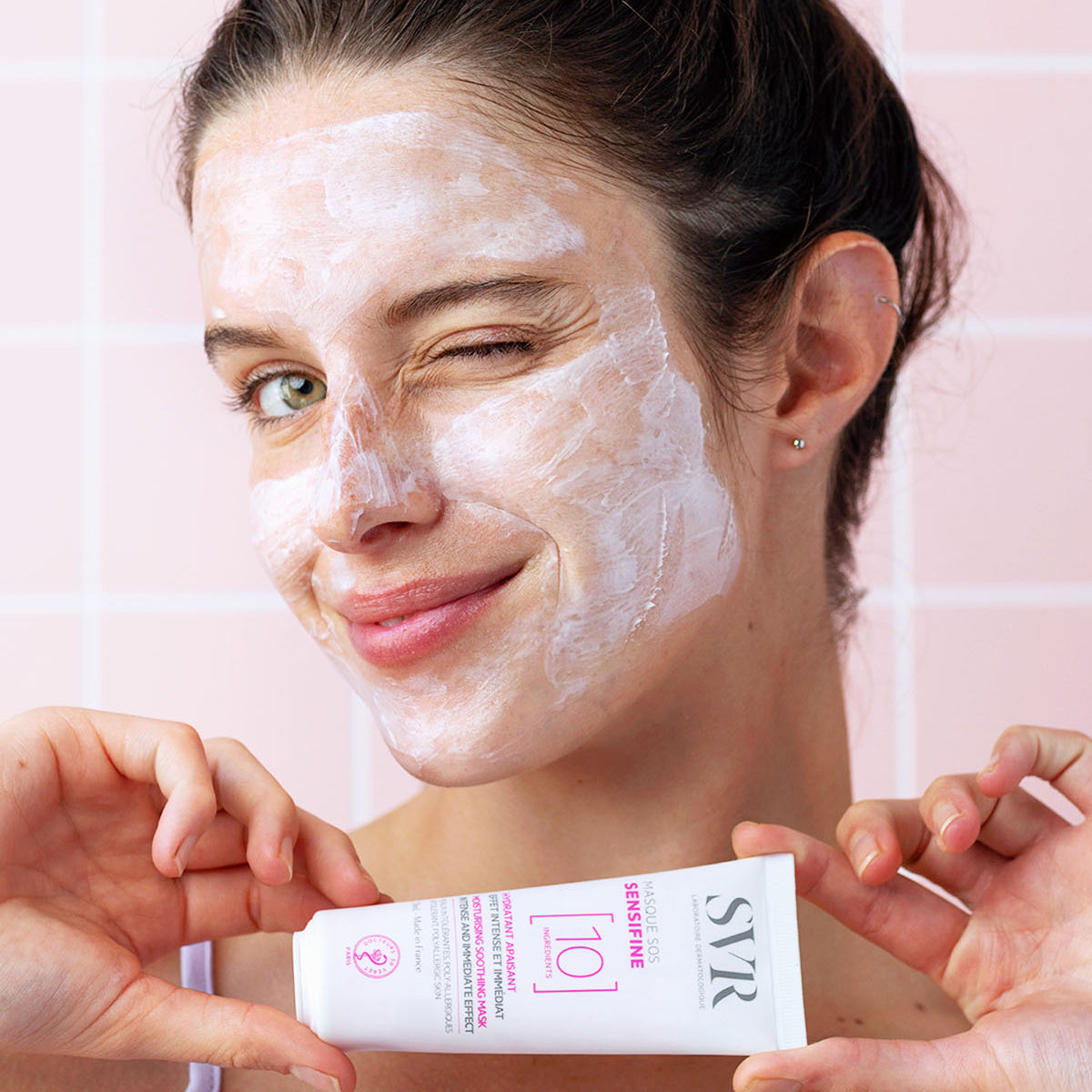 Femme souriante avec un masque SVR Sensifine rose sur le visage, tenant le tube blanc du produit devant un carrelage rose.
