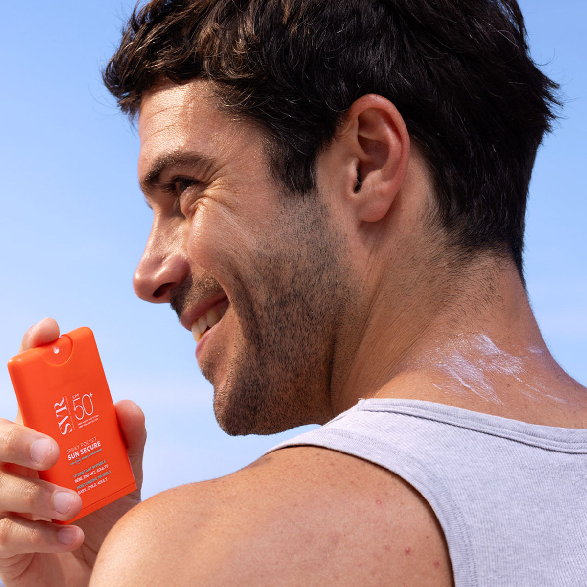Homme souriant tenant un spray solaire orange SVR Sun Secure SPF50+ format poche contre un ciel bleu.