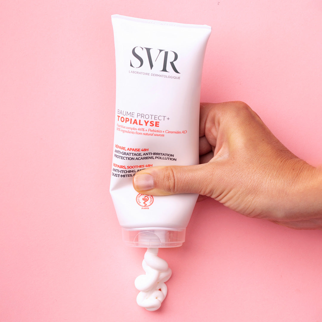 Main tenue pressant un tube blanc de baume SVR Topialyse Protect+ délivrant une texture crème onctueuse sur fond rose.