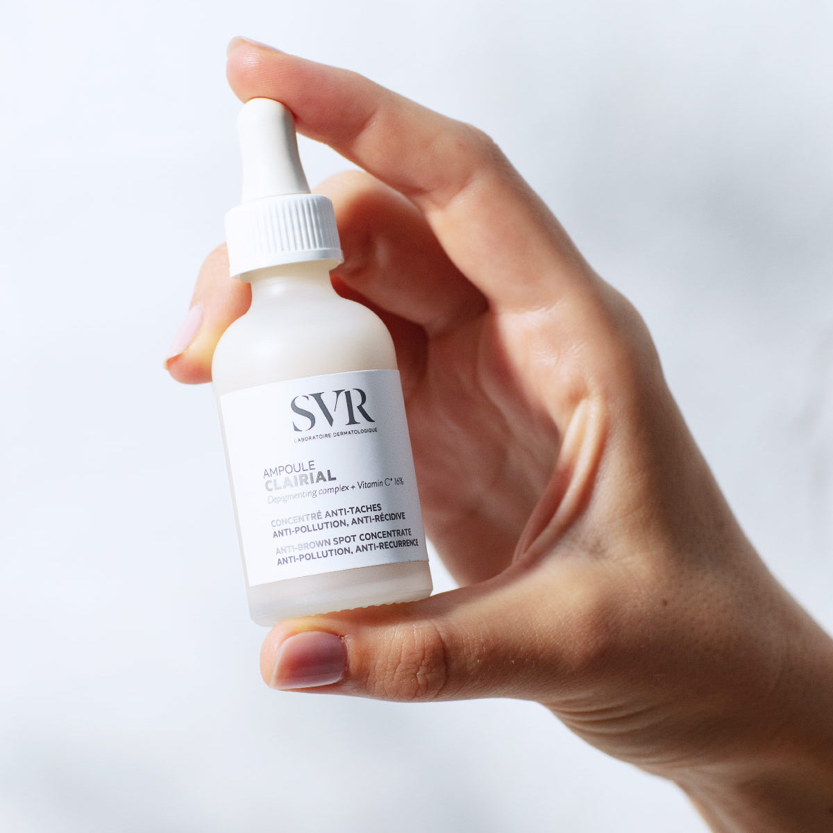 Main tenue à la main, flacon compte-gouttes en verre dépoli SVR Clairial Ampoule anti-taches avec étiquette blanche.