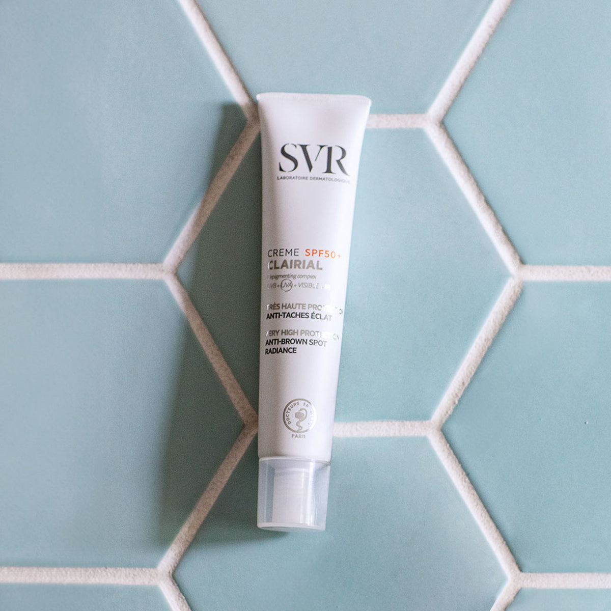 Tube blanc de crème SVR Clairial SPF50+ anti-taches sur un fond de carrelage hexagonal bleu clair.