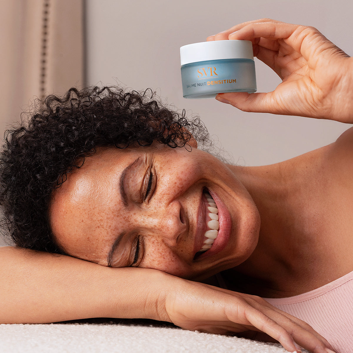 Smiling woman holding a frosted blue SVR Densitium Baume Nuit rechargeable jar with a white lid.