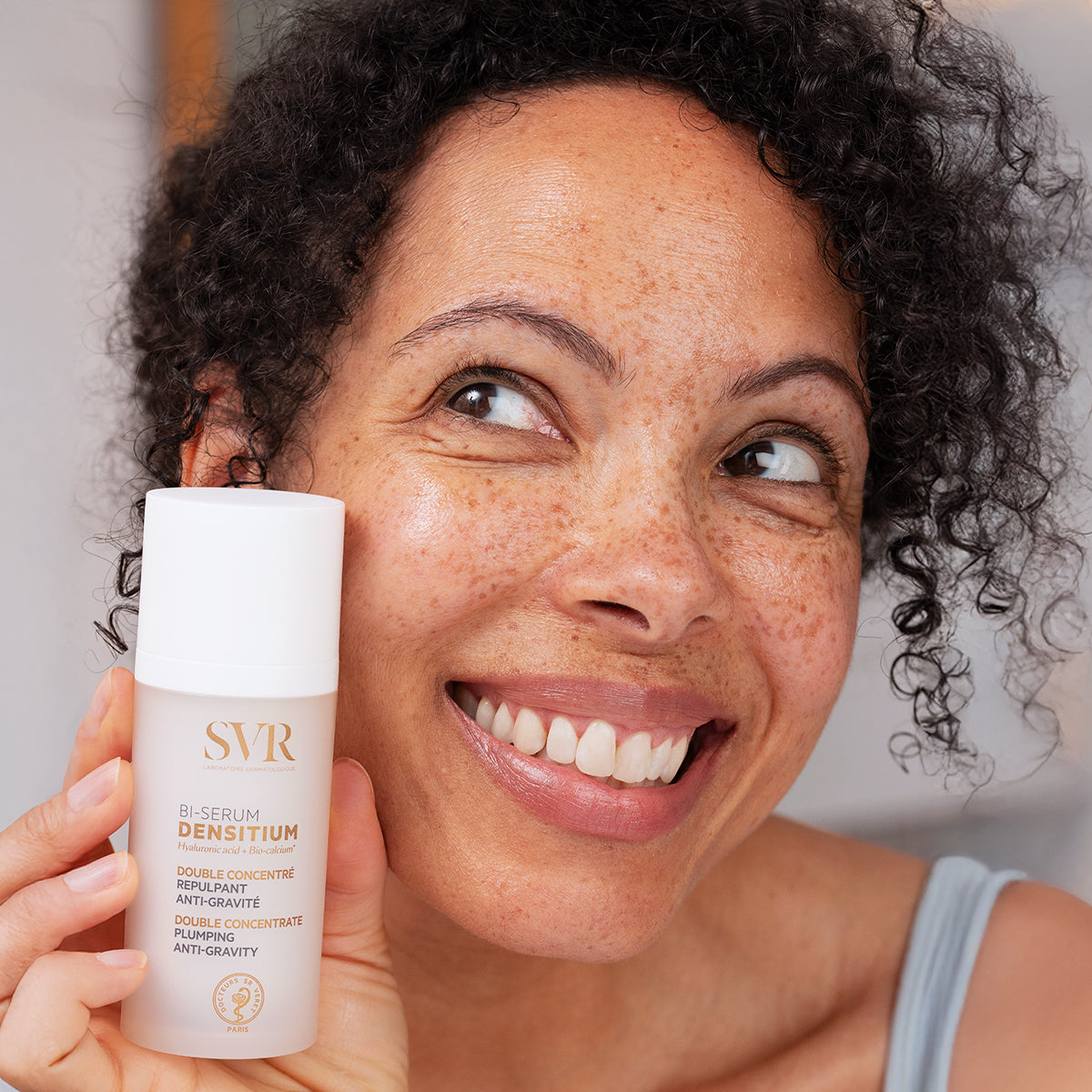 Smiling woman holding a white SVR Densitium Bi-Sérum bottle with gold text against a soft, neutral background.