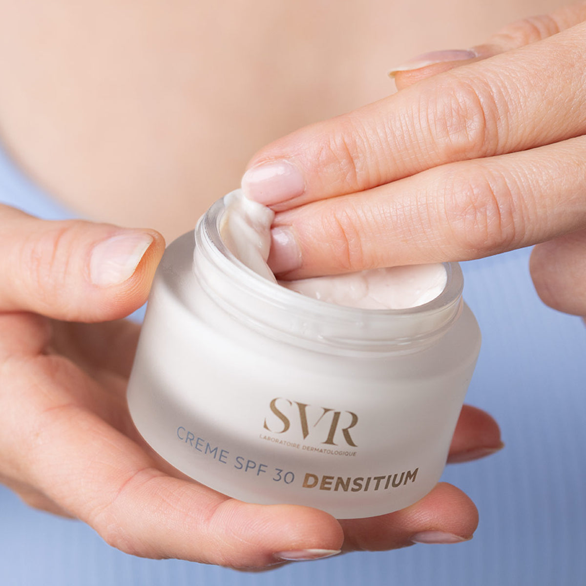 Hands holding a frosted glass jar of SVR Densitium SPF 30 cream, with a finger scooping the rich white face cream.