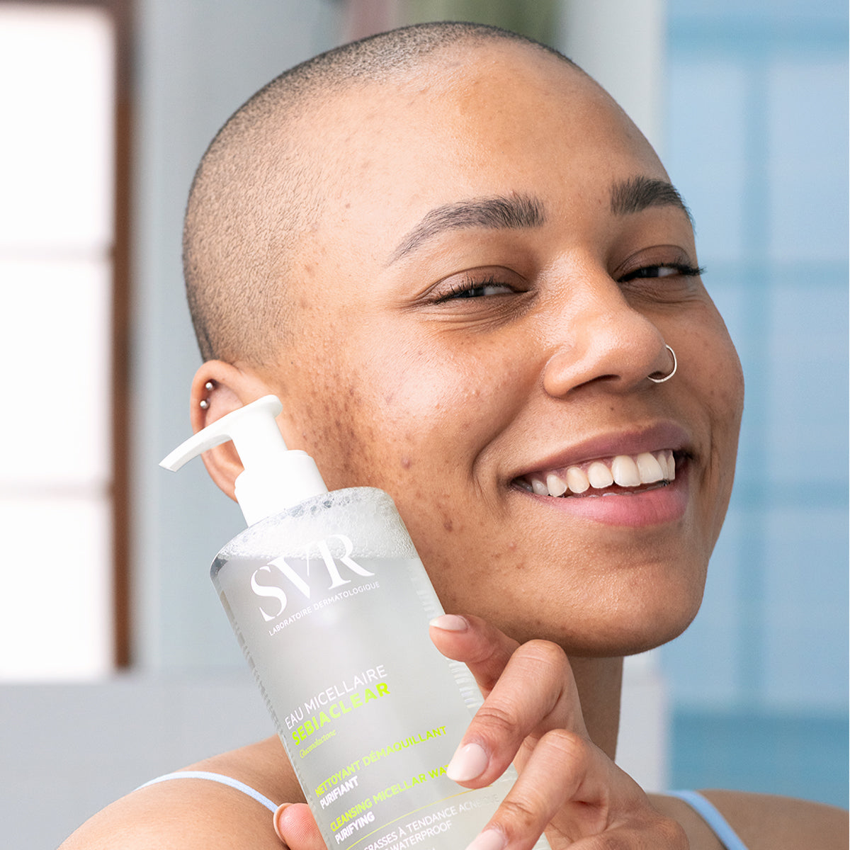 Femme souriante tenant un flacon pompe transparent d'eau micellaire SVR Sebiaclear pour peaux mixtes à grasses.