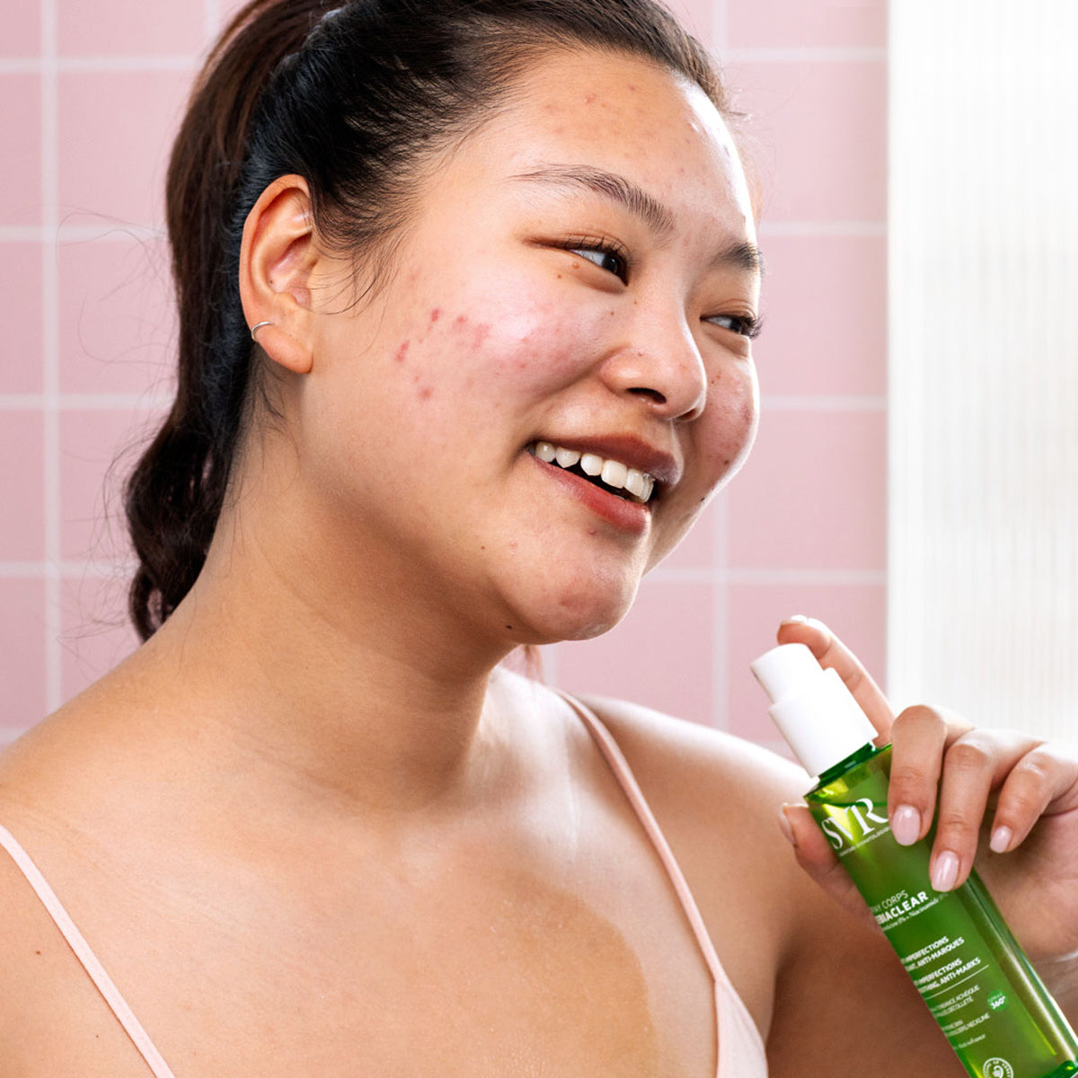 Femme souriante tenant un flacon spray vert SVR Sebiaclear Corps contre les imperfections sur fond de carrelage rose.