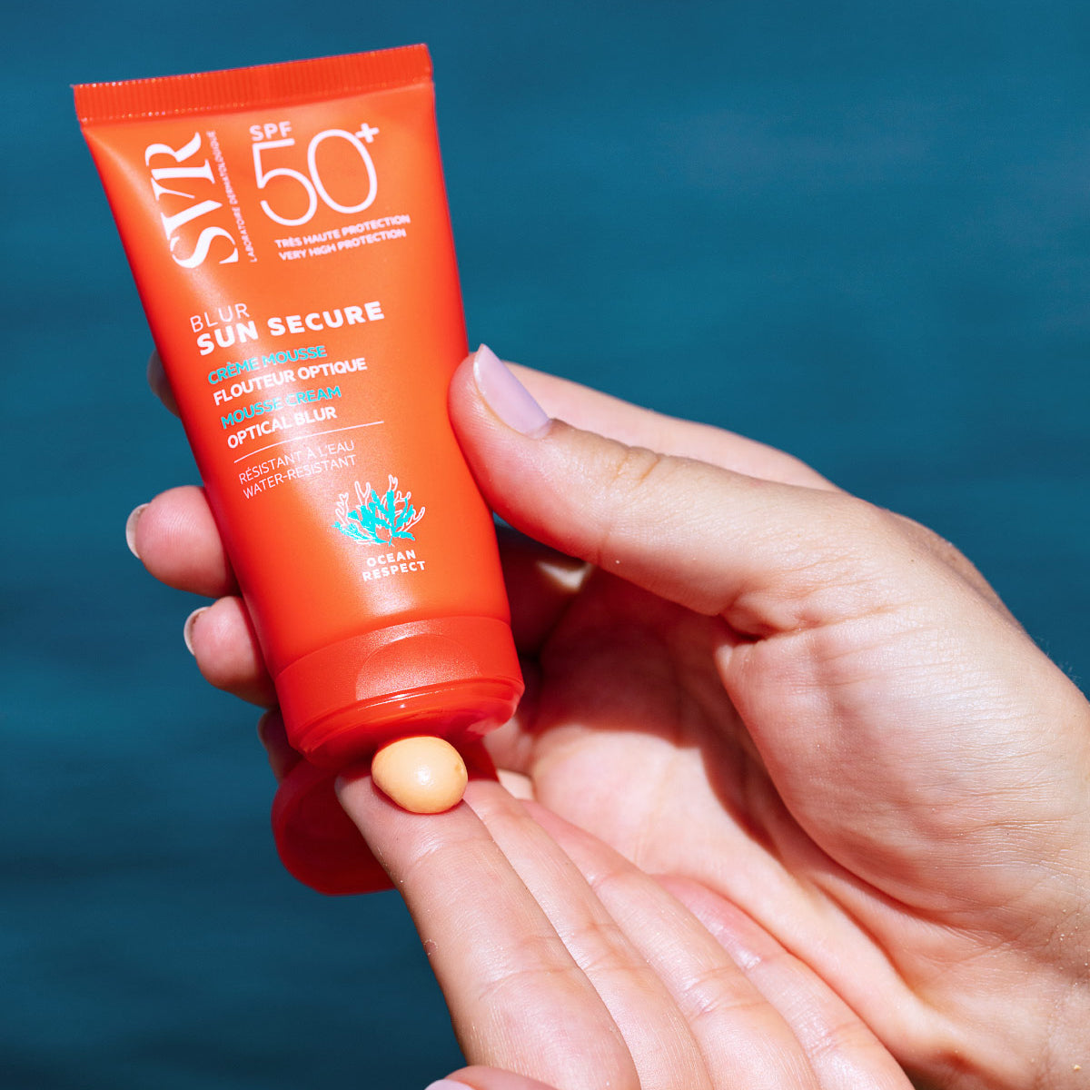 Tube orange SVR Sun Secure Blur SPF50+ avec une noisette de crème mousse abricotée sur le doigt, sur fond bleu océan.