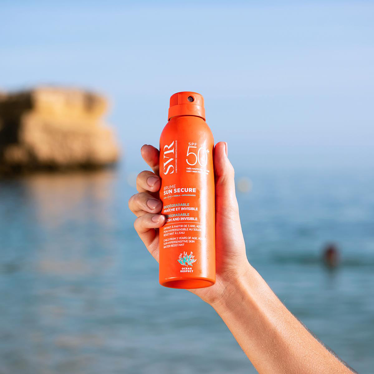 Main tenant un spray solaire orange SVR Sun Secure SPF50 devant une plage ensoleillée en arrière-plan.