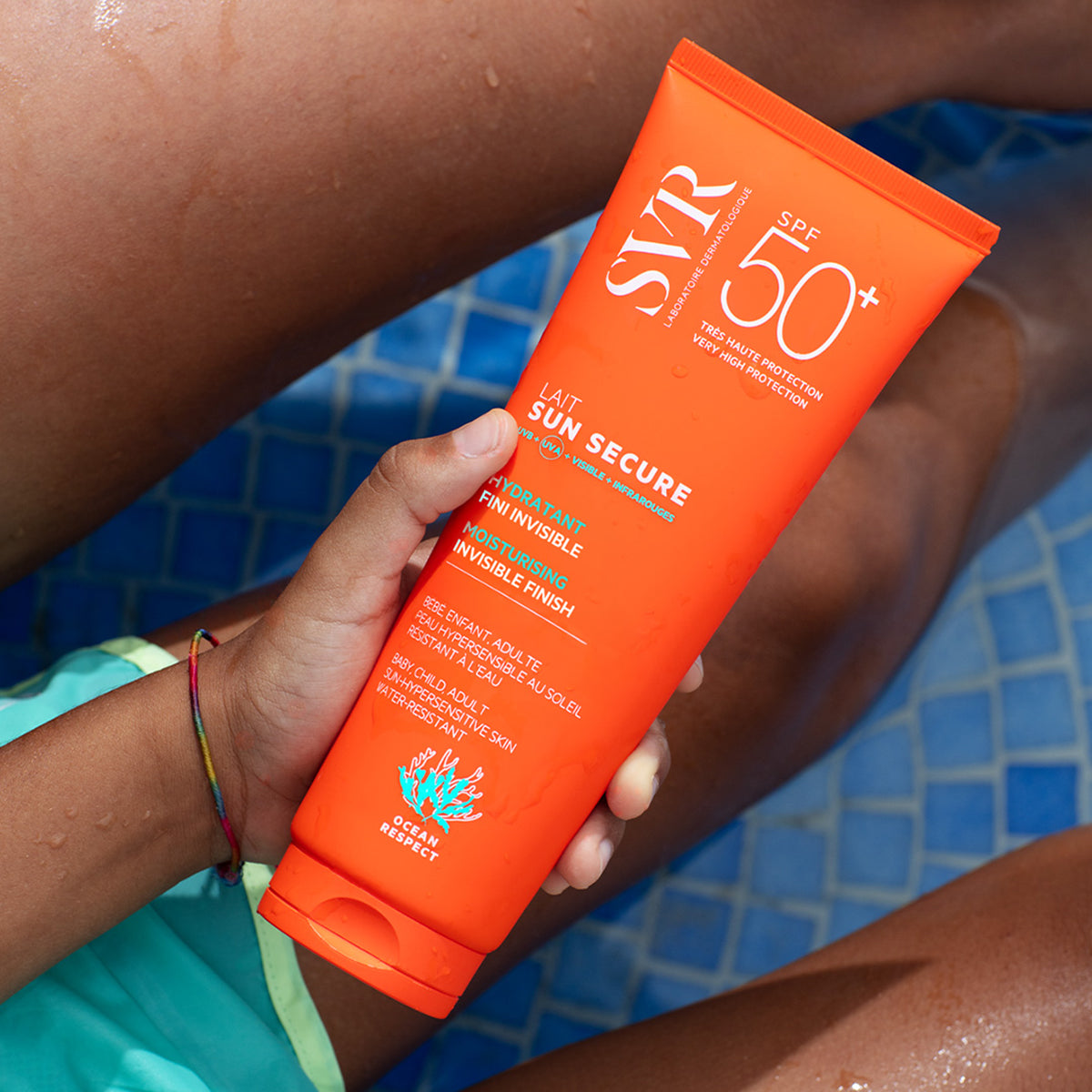 Tube orange de lait solaire SVR Sun Secure SPF50+ tenu à la main au bord d'une piscine bleue.